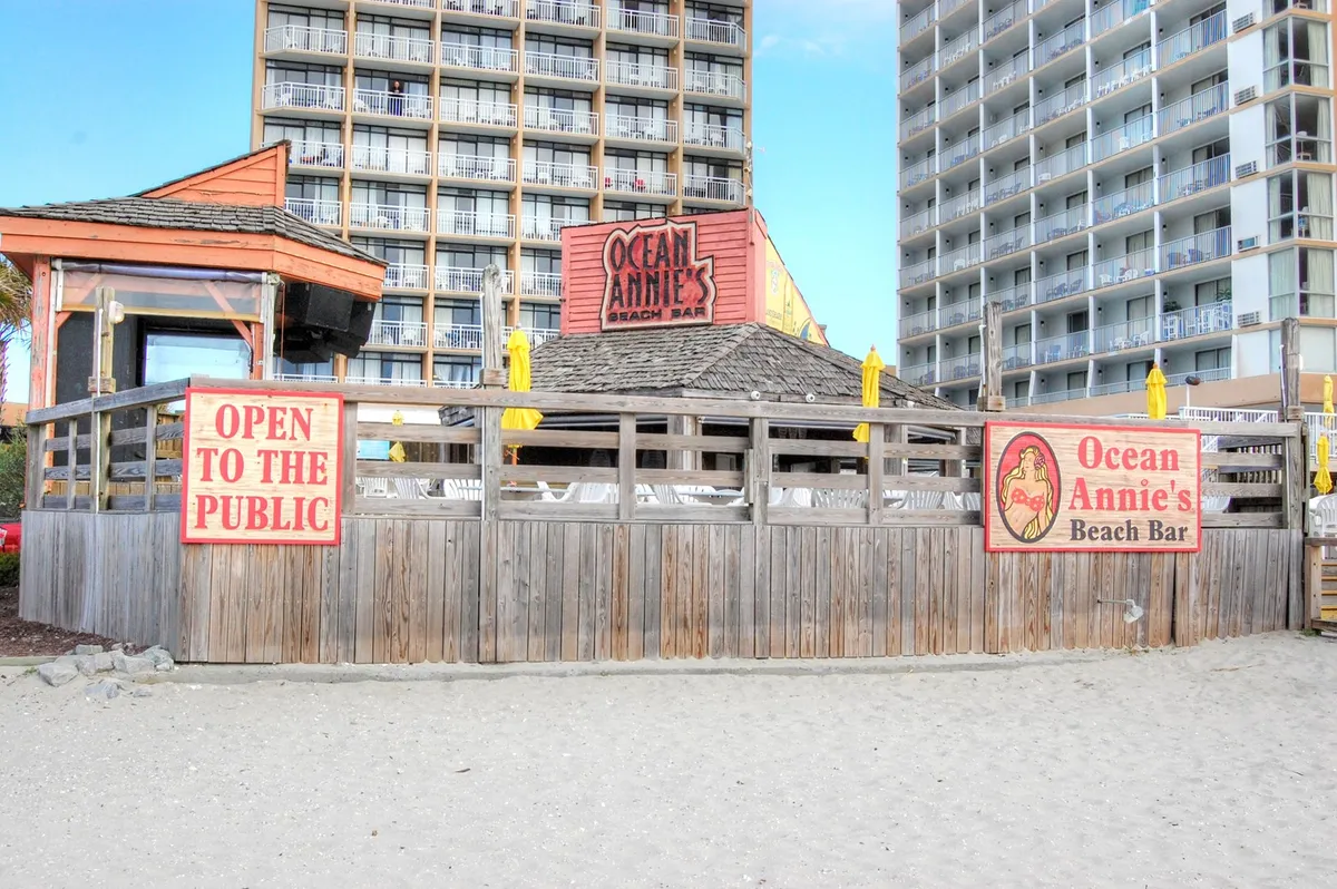 The photo shows the welcoming Ocean Annie's Beach Bar with its prominent signage and open layout. The casual, oceanfront setting is designed for guests to enjoy live music, tropical drinks, and relaxed socializing with the sound of waves in the background.