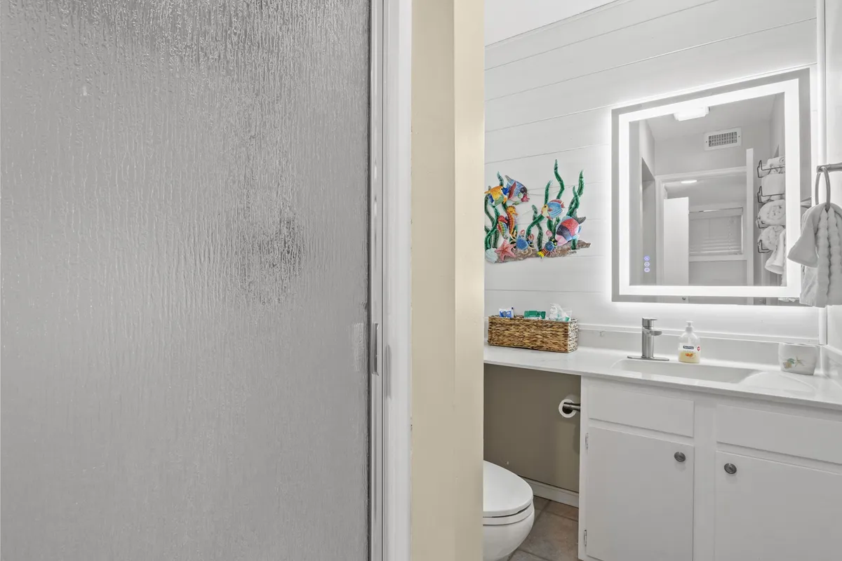 A modern bathroom with clean white cabinetry, a brightly illuminated mirror, and colorful wall art. The space feels fresh and cheerful, ideal for morning routines or relaxing after a long day.