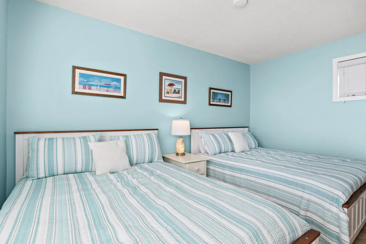 A bright and airy bedroom featuring two queen beds adorned with crisp linens and plush pillows. A nightstand with a soft-glowing lamp sits between the beds, and colorful framed artwork adds a touch of personality to the space.