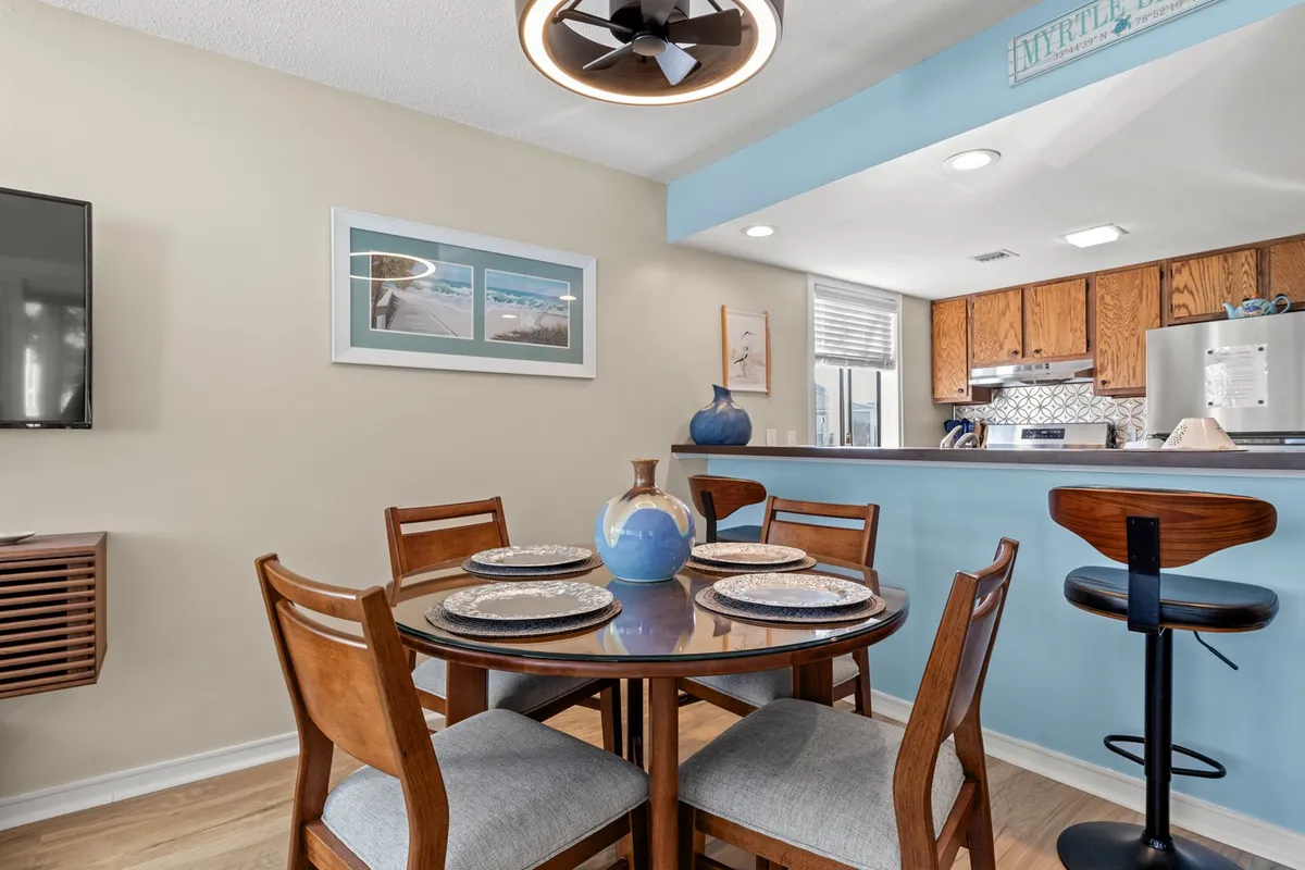An adjacent dining area with a round table and four chairs, a decorative blue vase, a wall-mounted TV, and a kitchen bar area in the background. This setting invites casual dining, family gatherings, and the enjoyment of meals together.