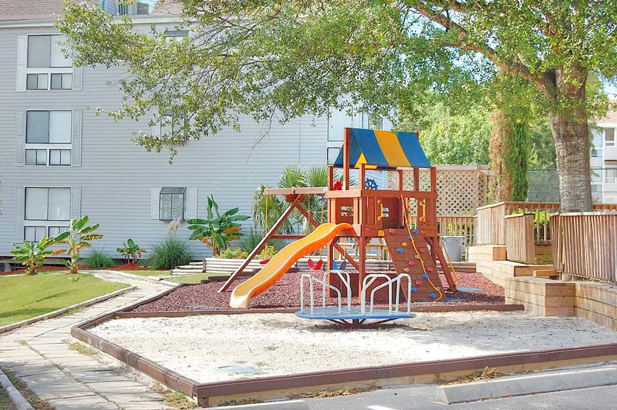 This exterior image showcases a vibrant playground area surrounded by lush greenery, with children playing and families gathering. The scene suggests community events, outdoor activities, and the welcoming neighborhood environment.