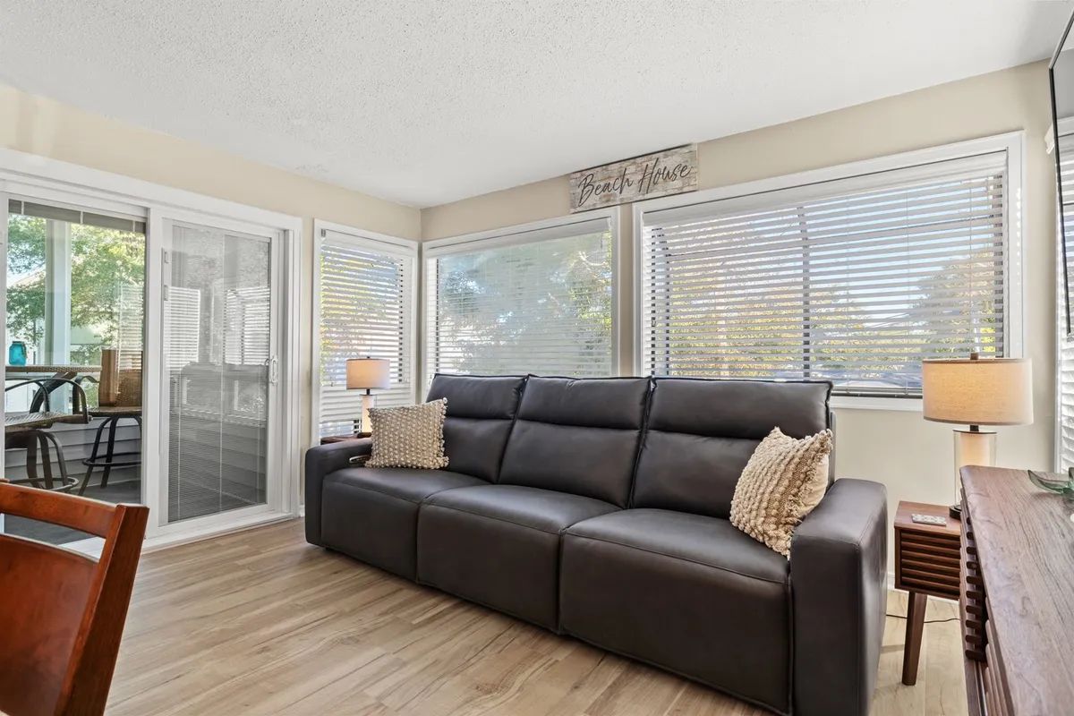 A leather sofa is framed by soft lighting from lamps and natural daylight streaming in through the windows. A beach house sign adds a coastal touch, and the arrangement is perfect for evening conversations or enjoying the morning sun, reinforcing the coastal theme.
