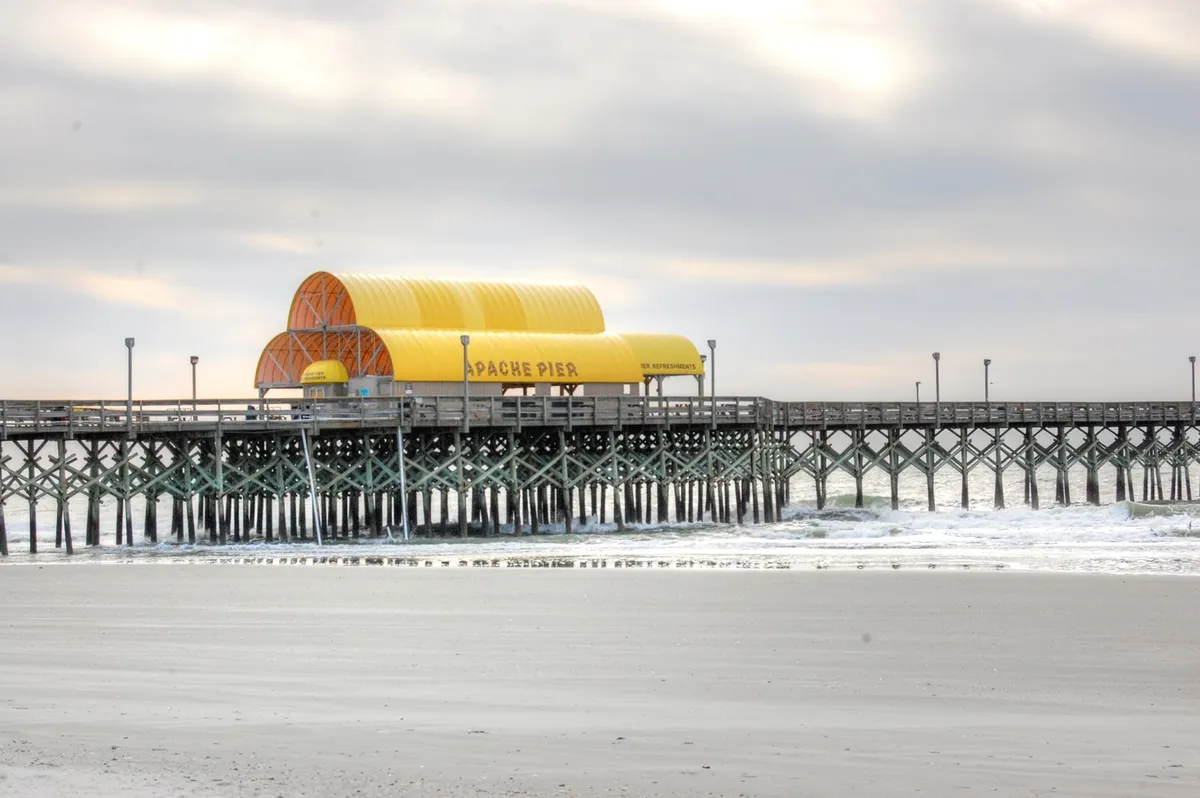 This image captures the iconic Apache Pier stretching out over the gentle waves, with its recognizable yellow roof and the shoreline in view. The serene ocean setting showcases the proximity to the beach, inviting visitors to fish, stroll, or simply enjoy the coastal atmosphere.