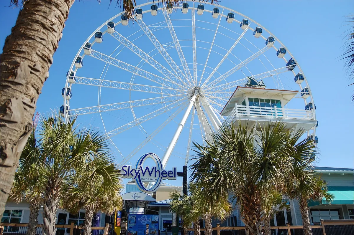 This image features the iconic SkyWheel, surrounded by palm trees and a ticket booth, set against a bright sky. The wheel towers above the boardwalk, inviting visitors to take a ride and experience panoramic views of the coastline. People are seen enjoying the lively, outdoor atmosphere.