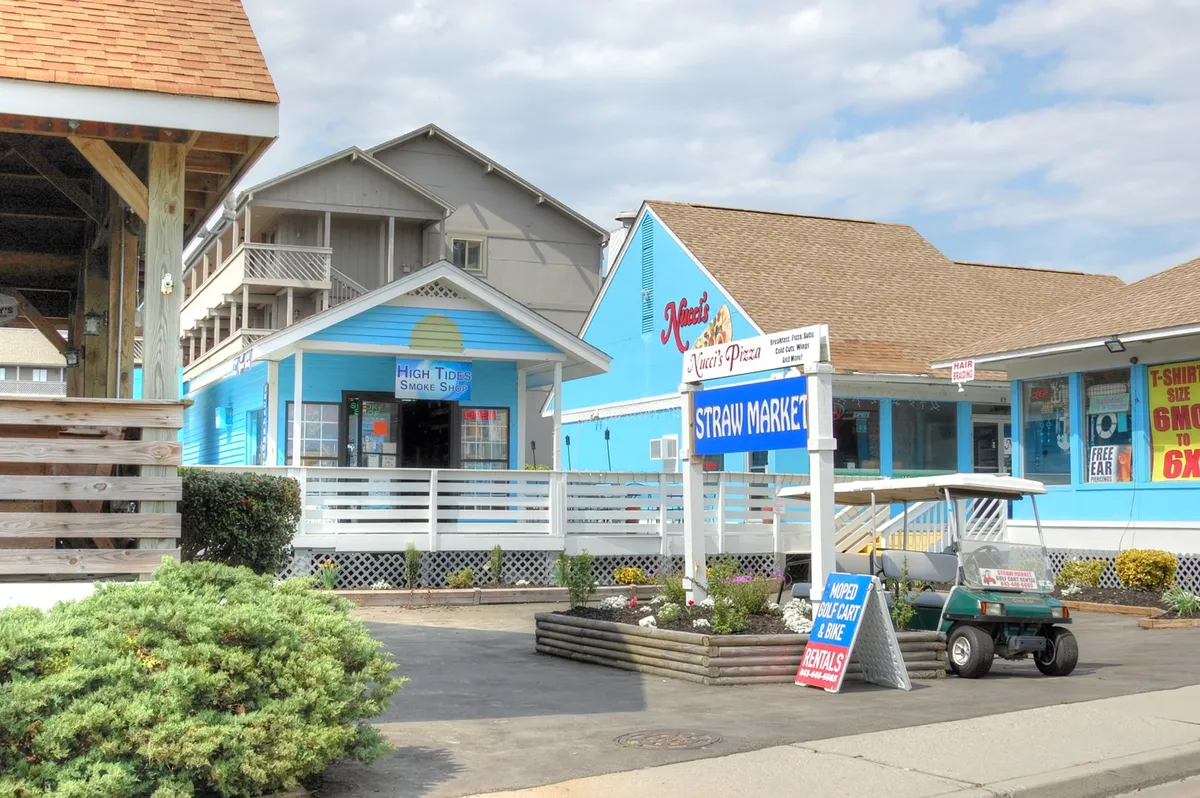 This image shows the Straw Market area with visible signage for local shops like 'Straw Market' and 'High Tide Smoke Shop,' set against charming blue buildings. The scene is bright and inviting, hinting at opportunities for shopping, dining, and leisurely strolls. People are likely enjoying the relaxed coastal vibe, making it a suitable backdrop for romantic and exploratory outings.