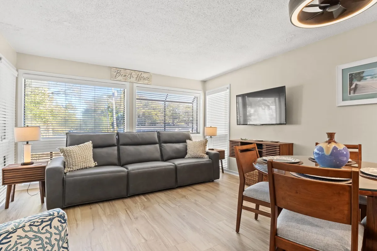 This image captures a well-lit living and dining area with a comfortable sofa, a spacious dining table, and large windows that allow abundant sunlight to pour in. The layout invites relaxation and socializing, with the windows creating a bright, airy atmosphere ideal for gatherings.