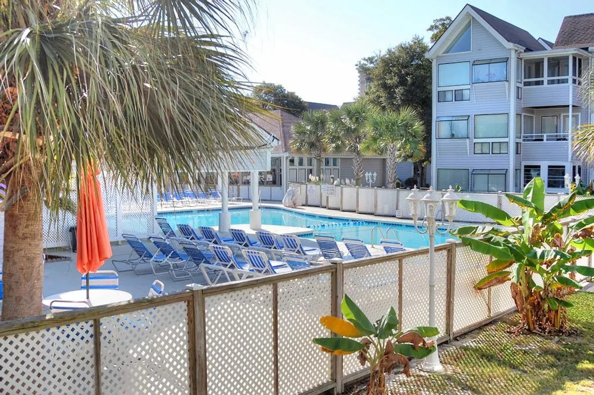 A sparkling swimming pool is surrounded by comfortable lounge chairs and shaded by palm trees and a bright orange umbrella. The relaxed atmosphere is ideal for sunbathing, swimming, and socializing with friends and family.