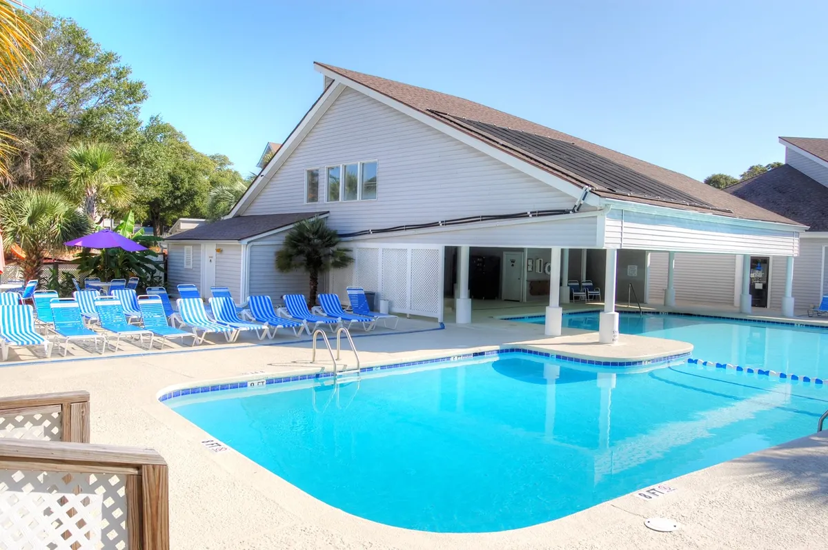 The image shows a sparkling outdoor swimming pool surrounded by inviting lounge chairs and a charming clubhouse, set against the backdrop of a relaxed and sunny Myrtle Beach atmosphere. The area is designed for relaxation, socializing, and enjoying the coastal vibe, with ample space for guests to swim, sunbathe, and gather with friends or family.