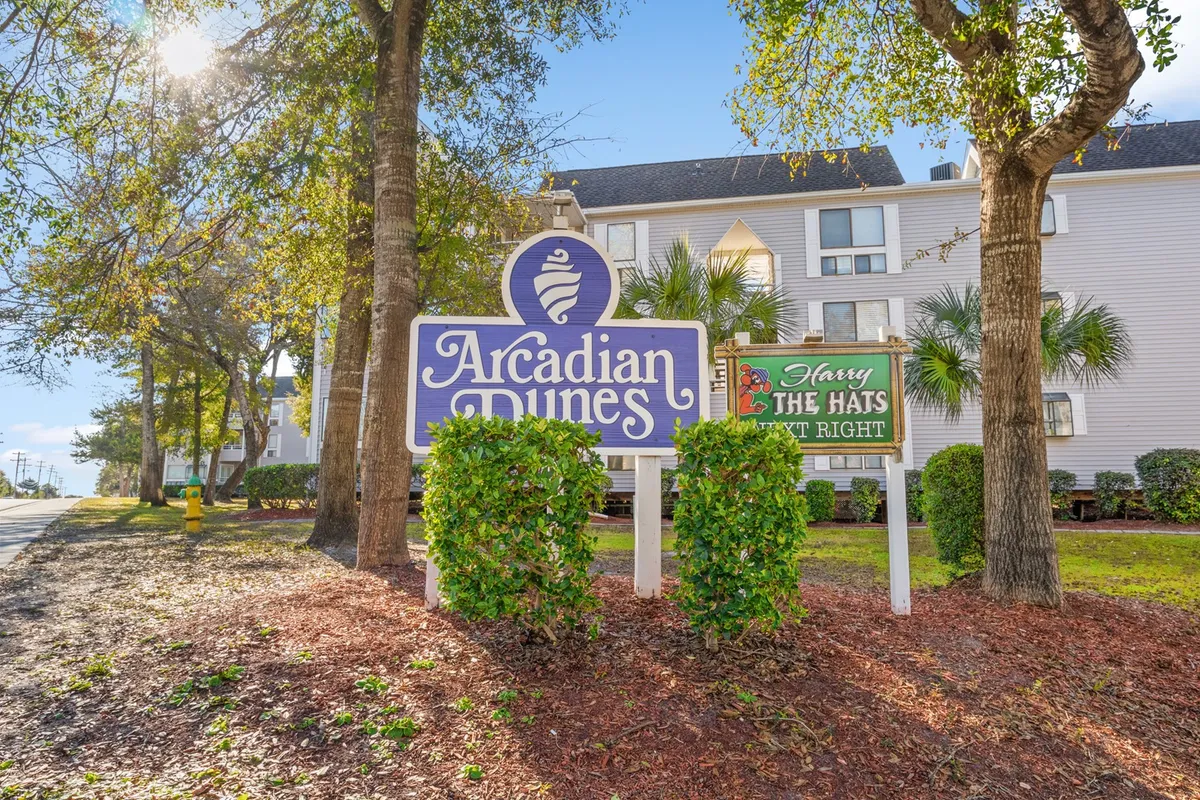The welcoming entrance to Arcadian Dunes, featuring the community sign and the Harry the Hats restaurant sign, is framed by lush greenery and palm trees, creating a cheerful and inviting atmosphere for arrivals and casual dining.