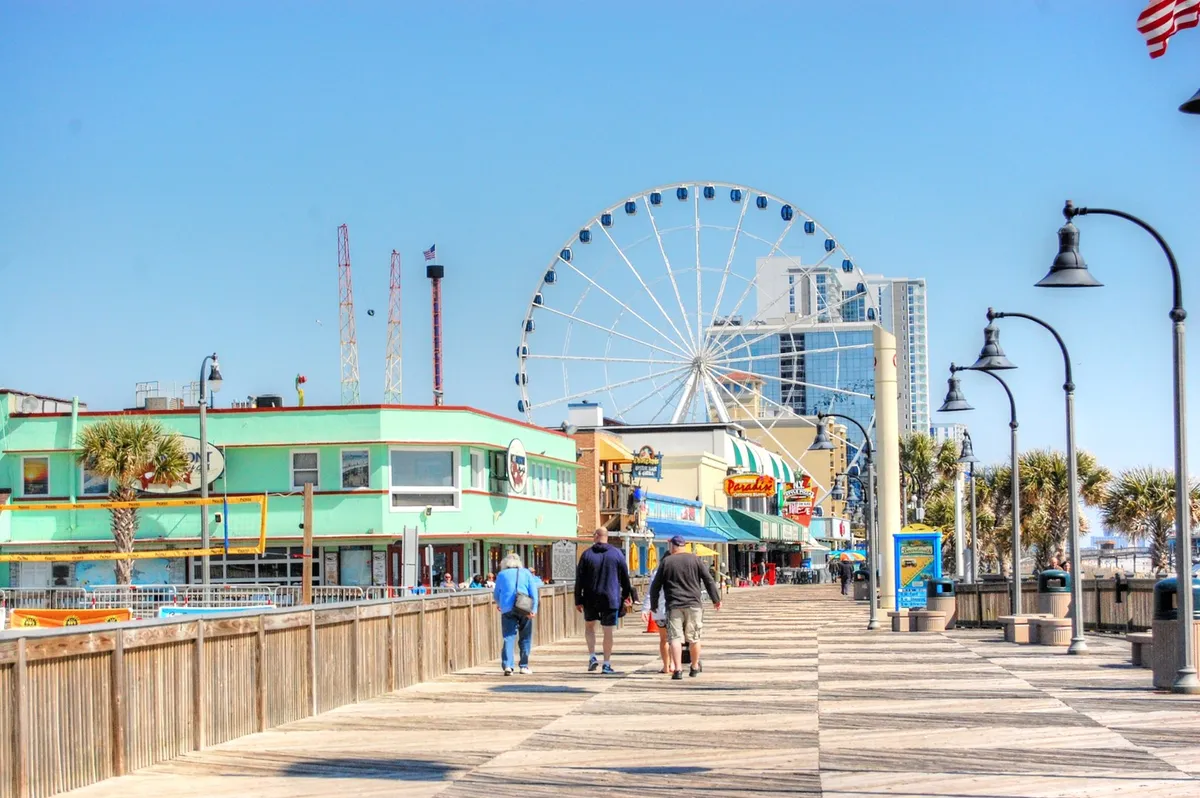 This image captures the vibrant Myrtle Beach boardwalk with a colorful ferris wheel, lively buildings, and people walking and enjoying the attractions. The scene radiates energy, fun, and the spirit of a classic family outing.