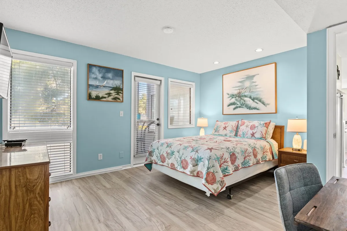 A serene bedroom with a single queen bed dressed in floral bedding. The room is filled with soft natural light from the large windows, features soothing artwork, and includes a sturdy wooden dresser for storage.